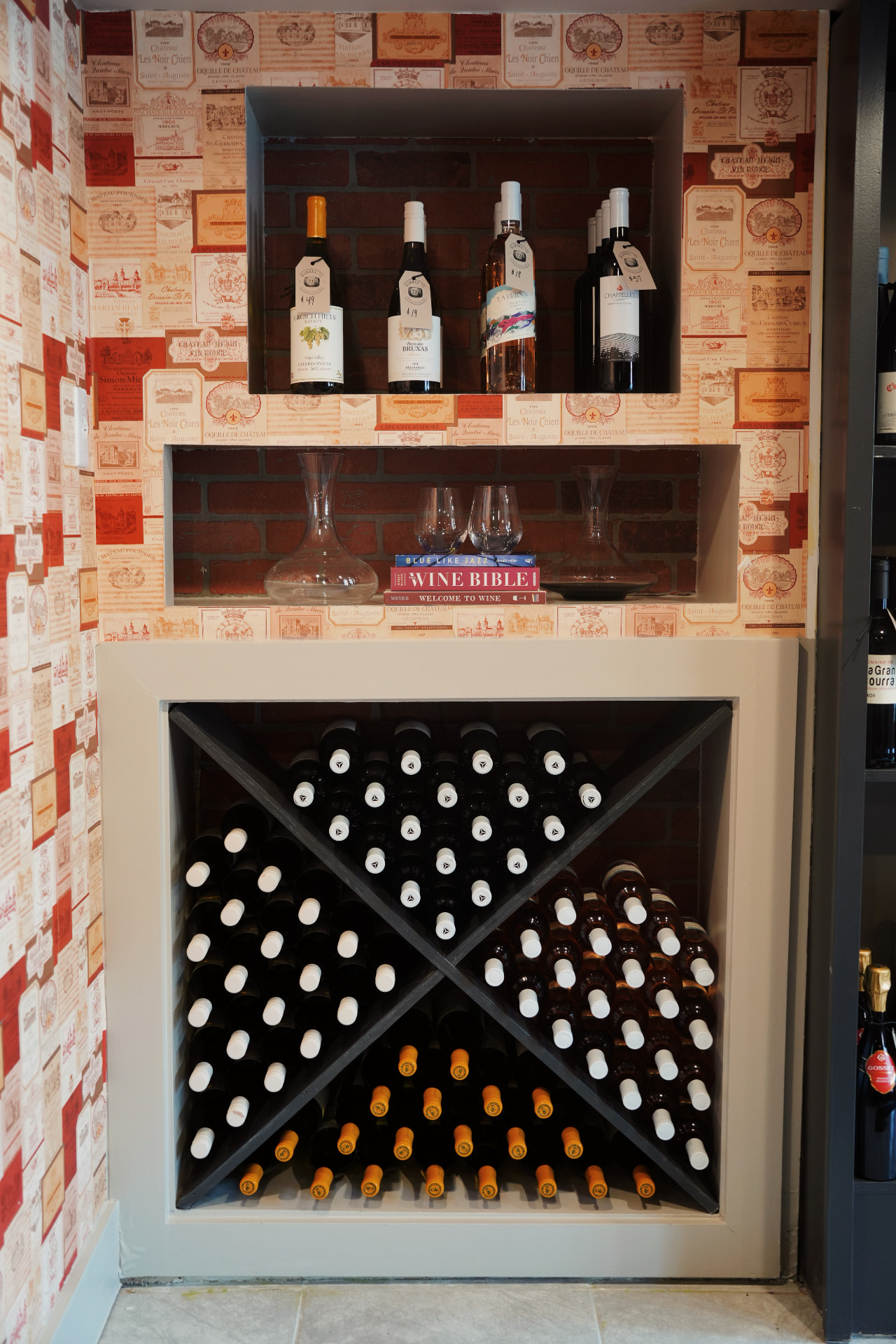 A wine alcove with bottles on display and a diamond-shaped rack full of bottles, set in wine-label wallpaper