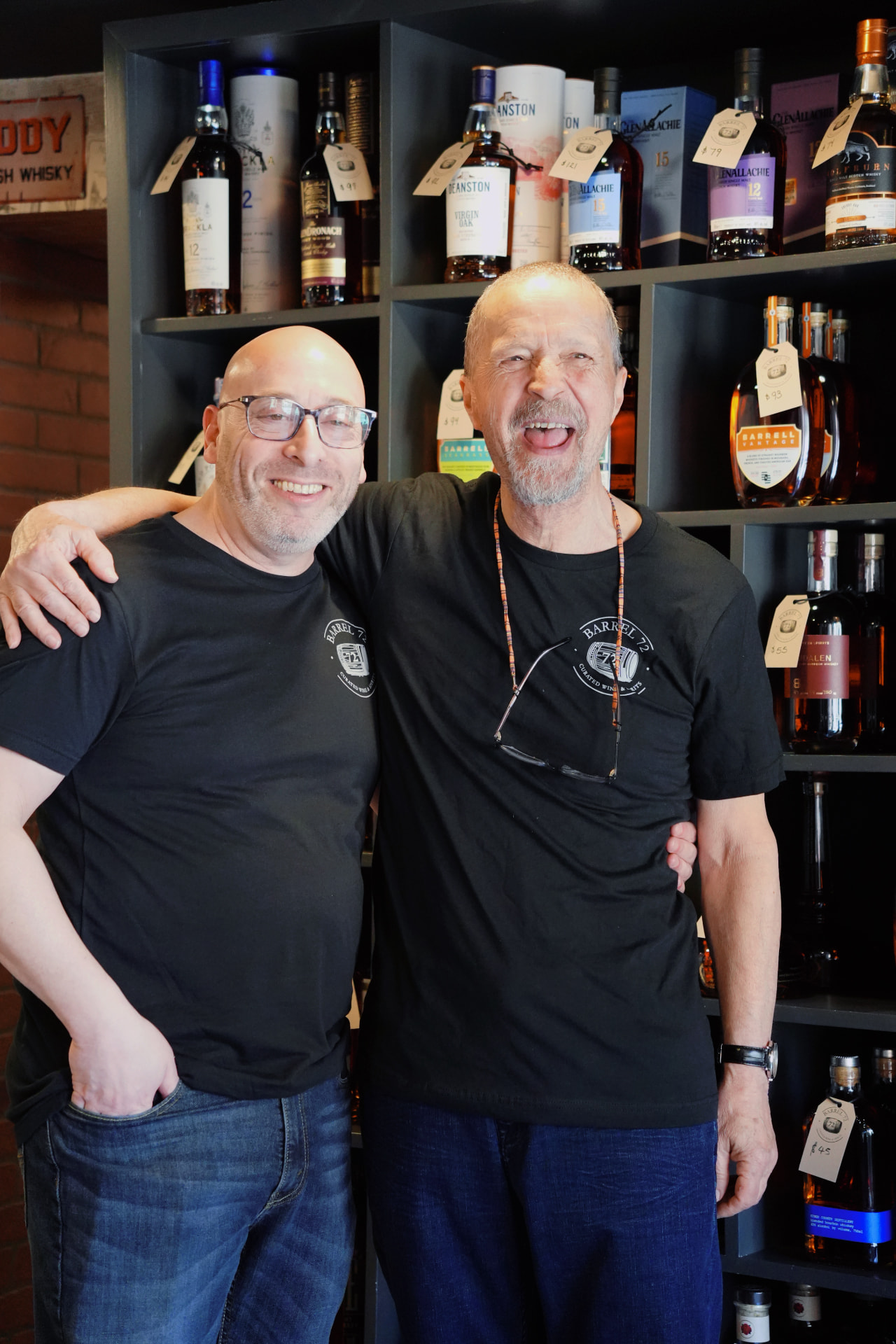 Two Barrel 72 team members in black shop shirts laughing arm in arm by the whiskey shelves