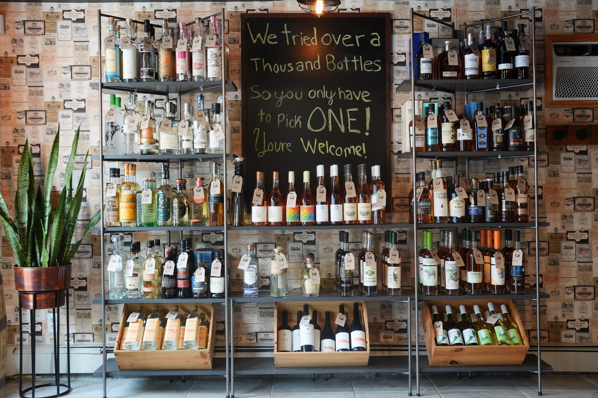 Metal shelves of wine and spirits around a chalkboard reading We tried over a thousand bottles, so you only have to pick ONE