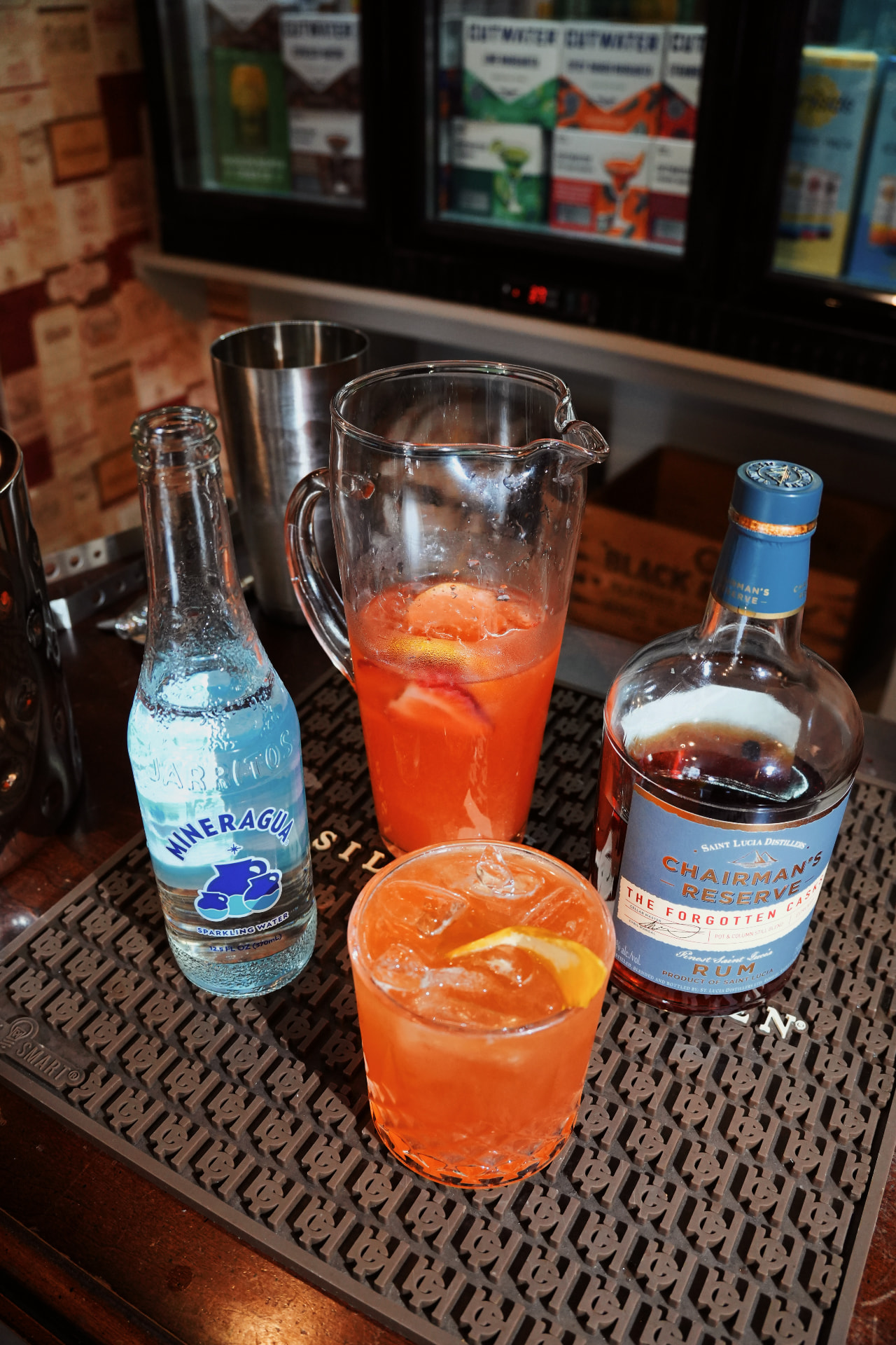 A pitcher and glass of pink rum punch next to a bottle of Chairman's Reserve rum and a sparkling water on a bar mat