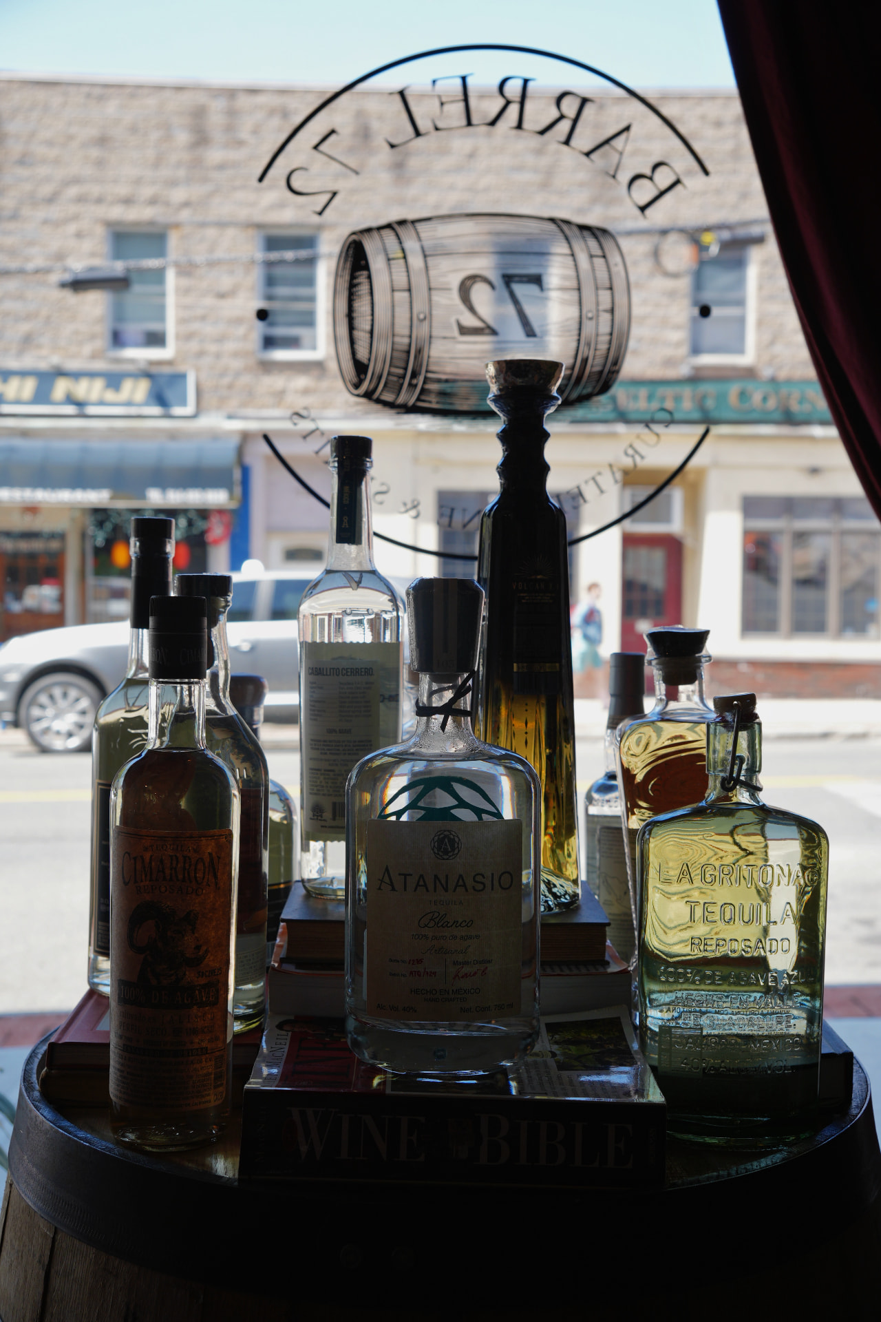 Tequila bottles, including Atanasio and La Gritona, displayed on a barrel in the Barrel 72 front window
