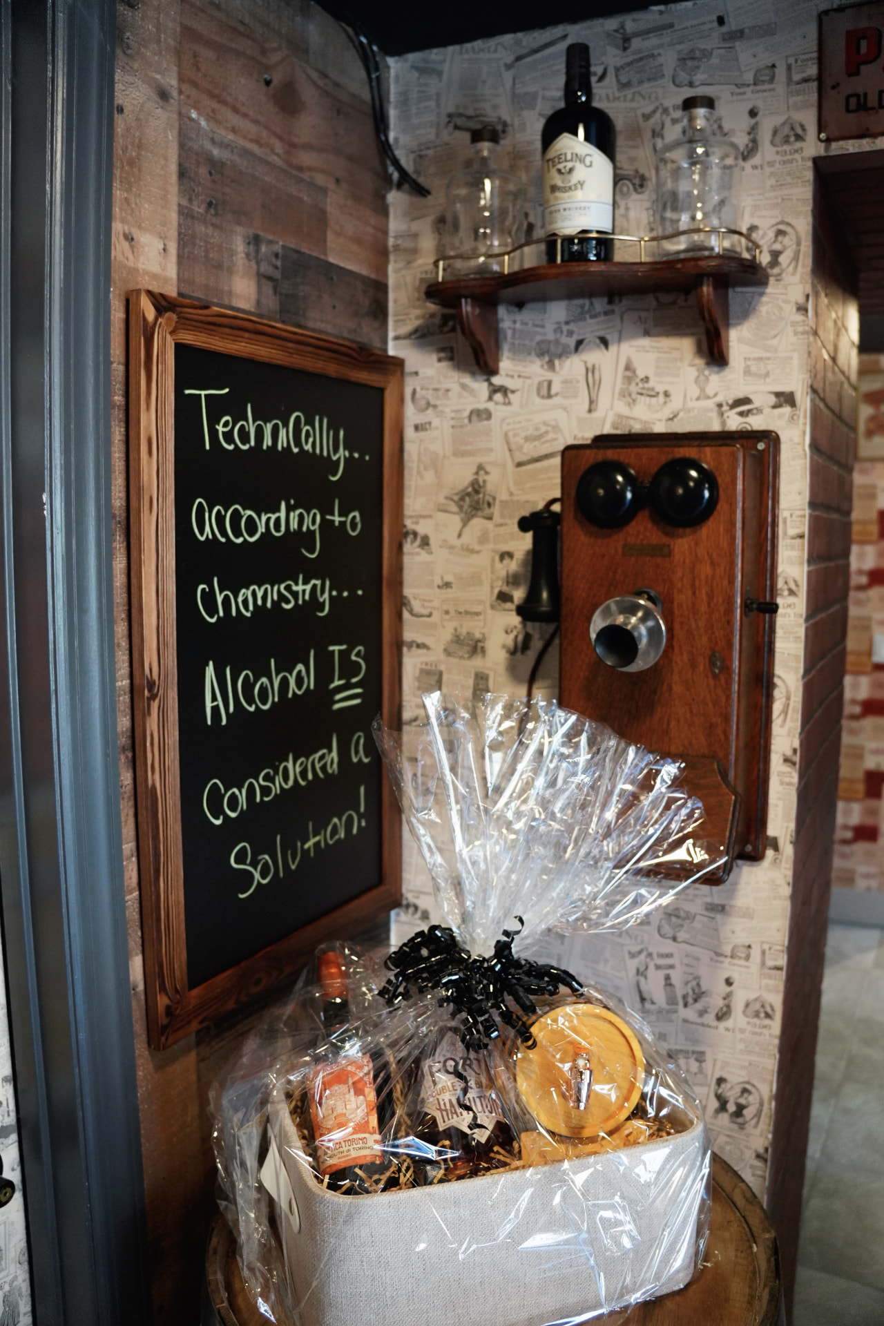 A cellophane-wrapped gift basket below an antique wall phone and a chalkboard that reads Technically, according to chemistry, alcohol is considered a solution